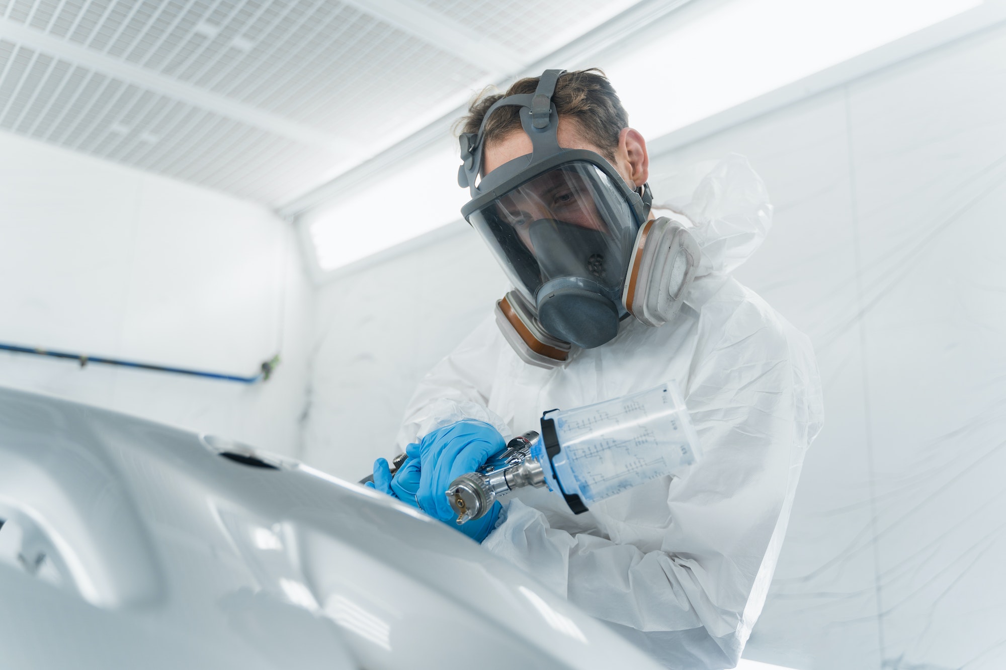 Car painter in protective clothes and mask painting and varnish automobile element in chamber